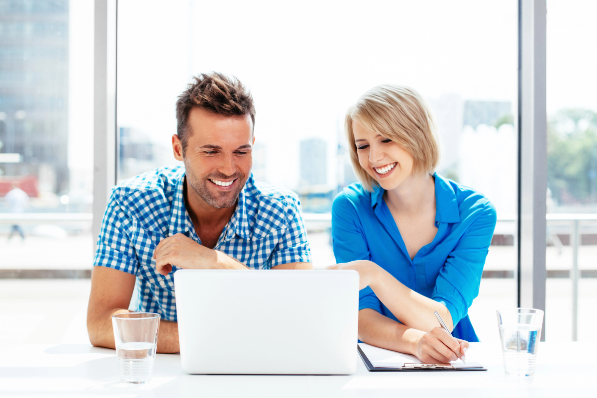 Two people smiling while looking at a laptop together in a bright office